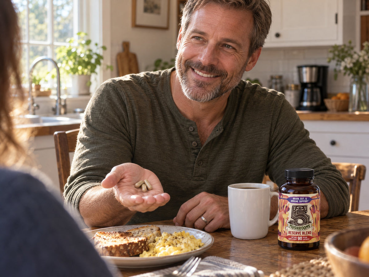 A man at breakfast holds three capsules next to the 5 Mushrooms bottle